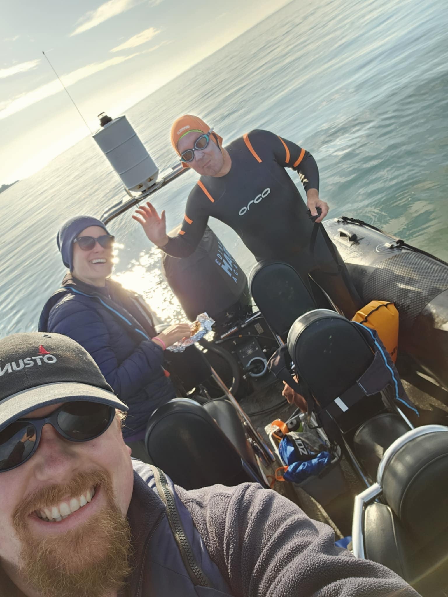 Three people are in a rubber dingy at sea. They are all wearing winter clothes and sun glasses and smiling. They feature a...